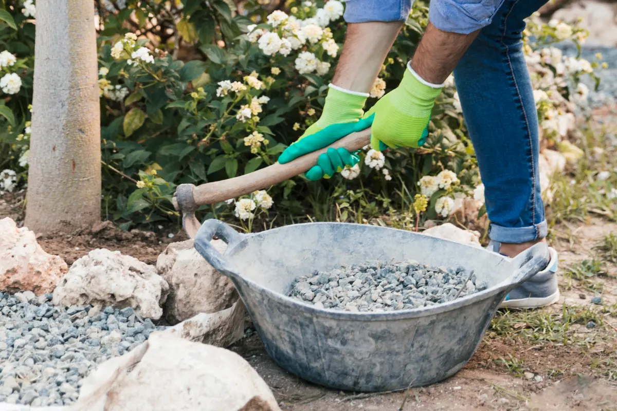 A worker uses a jackhammer to break up a cracked and damaged concrete driveway. The worker wears safety gear, including a hard hat, safety glasses, and ear protection. The driveway is surrounded by a lawn and landscaping.